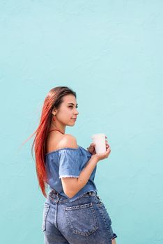 Attractive young woman in summer clothes and sunglasses holding cup of coffee in her hands, dancing on blue wall background at street