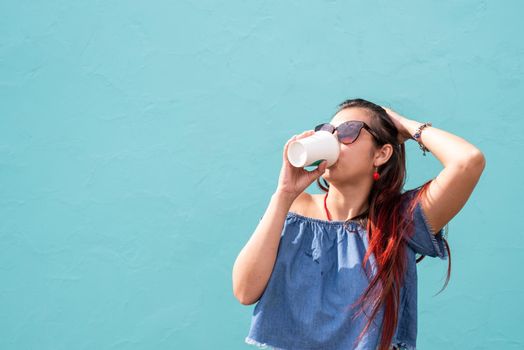 Attractive young woman in summer clothes and sunglasses holding cup of coffee in her hands, dancing on blue wall background at street