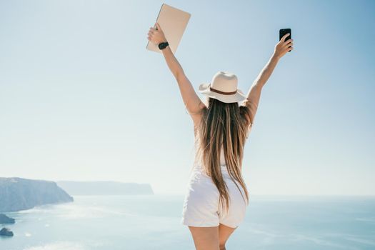 Successful business woman in yellow hat working on laptop by the sea. Pretty lady typing on computer at summer day outdoors. Freelance, travel and holidays concept.