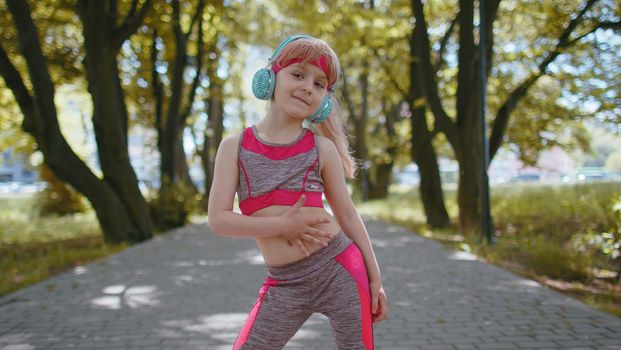Athletic fitness sport runner child girl training exercise, listening music on headphones dancing to camera having fun. Workout cardio in park. Toddler kid jogger enjoying funny dance. Active children