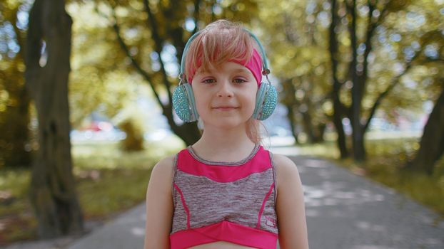 Portrait of athletic fitness sport runner child girl training, listening favorite music song in headphones. Workout cardio in morning park. Toddler kid jogger enjoy healthy lifestyle. Active children