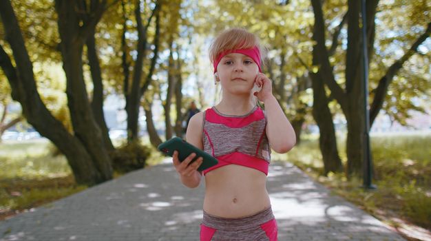Athletic fitness sport runner child girl walking and using smartphone listening music with wireless earphones. Workout cardio in morning park. Children toddler kid jogger enjoying healthy lifestyle