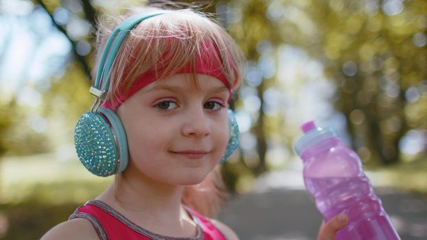Athletic thirsty sport jogger child girl drinking water from bottle after training exercising. Workout cardio in park at morning. Toddler kid runner enjoy healthy lifestyle. Active children activities