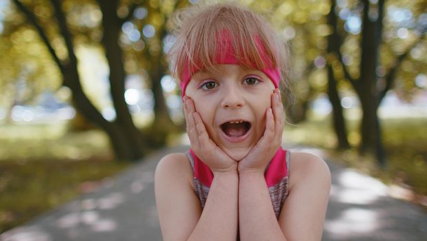Oh my God, Wow. Athletic fitness child kid runner girl raising hands in surprise looking at camera with big eyes, shocked by sudden victory. Workout cardio training outside in park. Active toddler