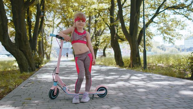 Athletic fit young girl rides on electric scooter on road in park on sunny day, outdoors. Child going standing near bike looking at camera. Kid leisure activity, sport. Modern technology for children