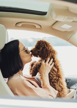 Young brunette woman happy owner of redhead dog toy poodle, sitting inside car and kissing her four paws pet