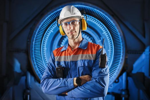 Portrait of a young professional engineer standing next to a gas turbine engine in a protective helmet, headphones and glasses. In the background is an unfocused industrial disk of a turbine rotor with blades.