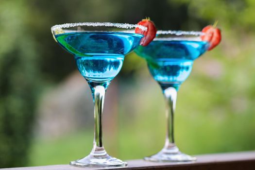 Two glasses with blue margarita cocktail garnished with lime zest and strawberries, selective focus.