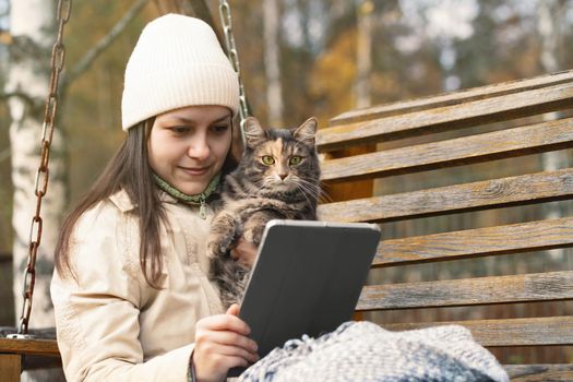 Girl uses a tablet while sitting on a bench in an autumn park with a cat in her arms.