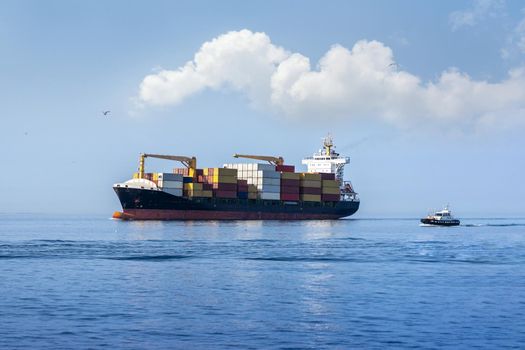 A fully loaded large cargo container ship at sea heading to port with the help of a small tugboat