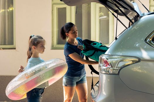 Mother with kid loading baggage in trunk, preparing to leave on road trip journey. Woman and small child travelling on holiday vacation at seaside during summer, happy recreation.