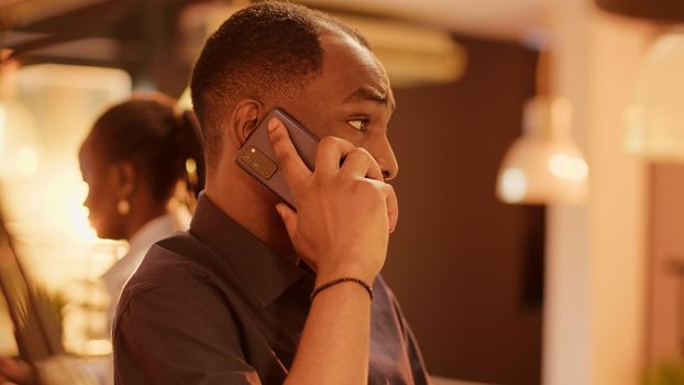 African american man talking on mobile phone call at office desk, using online phone line communication to chat remotely. Having remote conversation on smartphone, dial number.