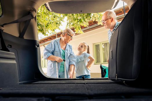 Grandparents and niece leaving on vacation, loading travel bags and baggage in automobile trunk. Senior people and little kid travelling on summer holiday road trip with vehicle, drive journey.
