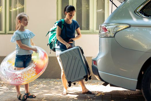 Mother and child travelling on vacation, loading baggage trolley and inflatable in vehicle trunk to go to seaside destination during summer holiday. Woman and little girl leaving on road trip.