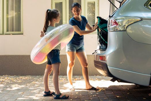 Mother with little girl loading baggage in trunk, preparing to leave on road trip journey. Woman and small child travelling on holiday vacation at seaside during summer, happy recreation.