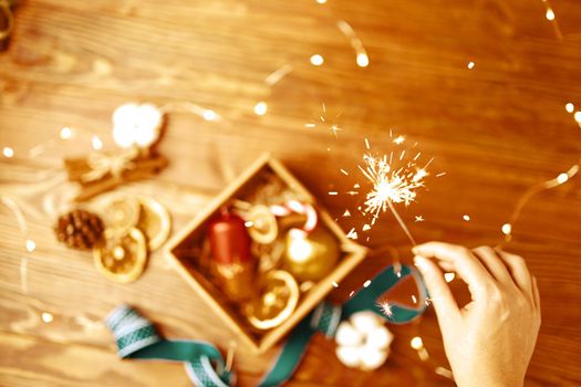 Woman's hand holds burning sparkler over wooden table with Christmas gifts. Box with ornate candle, golden ball, ribbon, dried oranges and cones. Festive composition for postcards.
