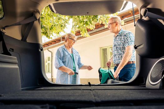 Elderly couple putting travel bags in trunk to leave on vacation journey with vehicle. People travelling together on retirement holiday trip with trolley and baggage, leisure activity.
