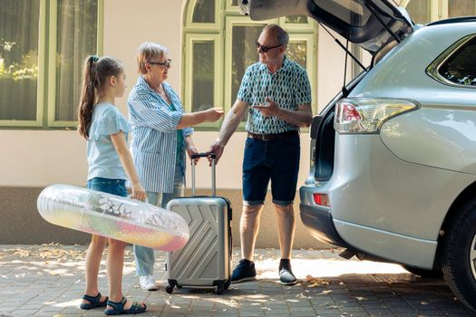 Grandparents and niece leaving on holiday, travelling on seaside vacation during summer. Senior couple and little girl going on journey trip with travel bags and inflatable in trunk of vehicle.