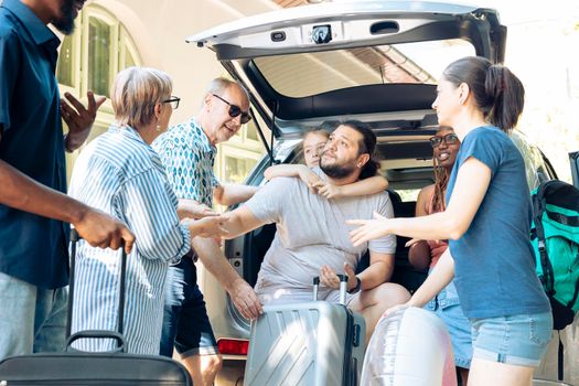 Mixed family and friends loading bags in automobile trunk, preparing to leave together on holiday trip. Travelling by car on summer vacation, going to seaside destination with multiethnic people.