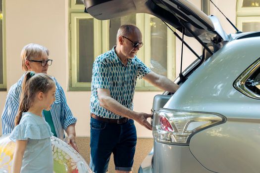 Senior people taking child on holiday by car, travelling on summer vacation at seaside. Grandparents and niece leaving on journey adventure, putting travel bags and inflatable in trunk.