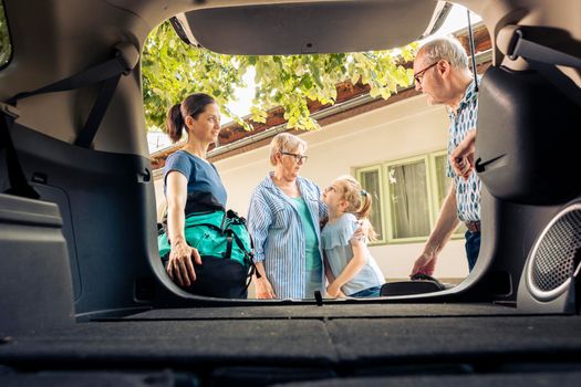 European family going on summer holiday with luggage and bags, loading baggage in automobile trunk. Little girl with mother and grandparents travelling on vacation trip with vehicle.