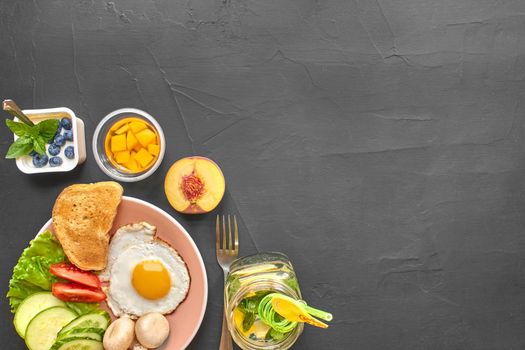 Top view of healthy and tasty meal on a black background with copy space. Fresh vegetables, scrambled eggs, toast are on a plate for delicious and nutritious breakfast. Yoghurt, peach, drink with green tubule are nearby.