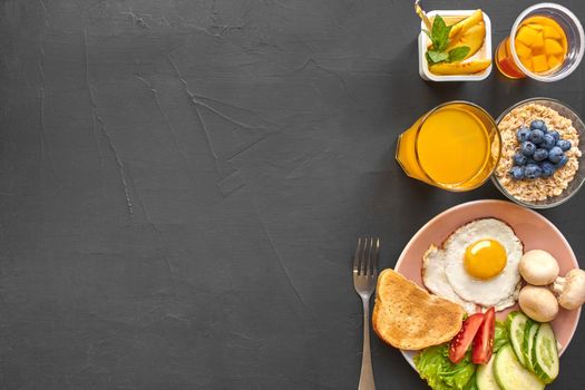 Top view of healthy and delicious breakfast on a black background with copy space. Fresh vegetables, scrambled eggs, toast are on a plate for tasty and nutritious meal. Oat flakes with a blueberry, yoghurt, orange juice and fork are nearby.