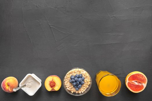 Top view of healthy and gourmet snack on a black background with copy space. Fresh fruits, oat flakes with blueberry, natural yoghurt and a glass of orange juice for tasty and nutritious meal.