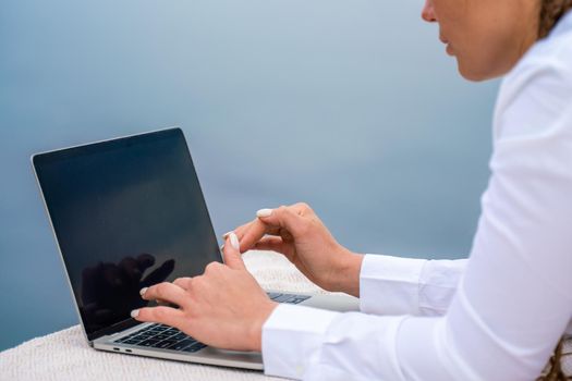 A woman is typing on a laptop keyboard on a terrace with a beautiful sea view. Close up of a woman's hands writing on a computer. Freelancing, digital nomad, travel and vacation concept