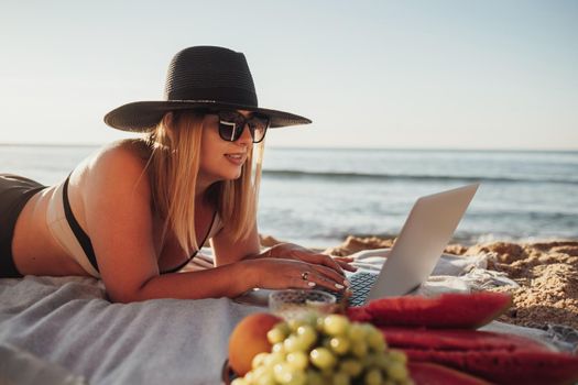 Pretty Young Woman in Sunglasses and Hat Using Laptop, Enjoying Picnic on Vacation While Lying on Sandy Beach by Sea