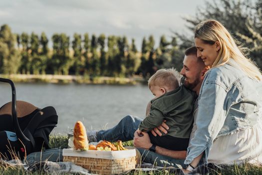 Happy Young Family, Mother and Father with Their Little Son Having Picnic Outdoors by Lake at Sunset