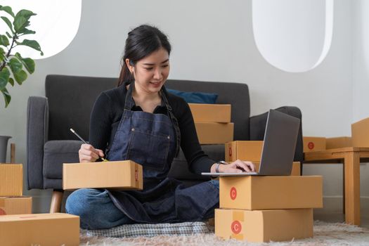 startup small business owner writing address on cardboard box at workplace. Freelance Asian woman small business entrepreneur SME working with box at home
