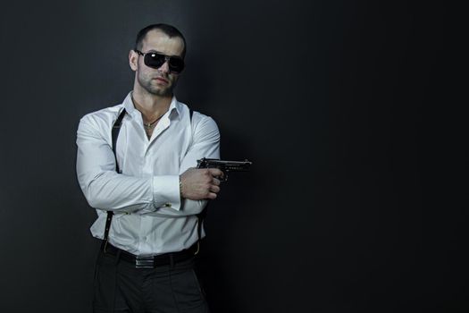 Young man in glasses wears a white shirt and suspenders, holds a gun in his hand