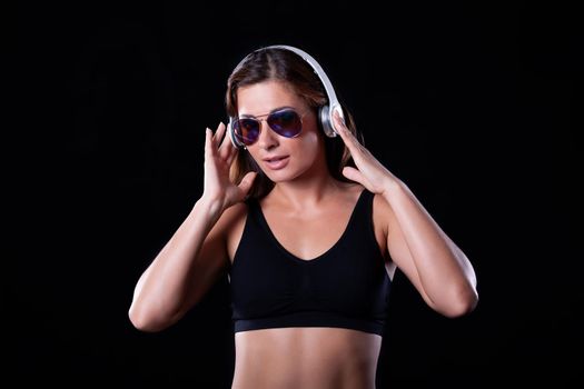 Sporty female brunette girl showing off her perfect body on black background with headphones and sunglasses. Close-Up.