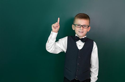 Happy cute clever boy in glasses raises a finger up. He had an idea. Child is ready to answer with a blackboard on a background. First time to school. Back to school.