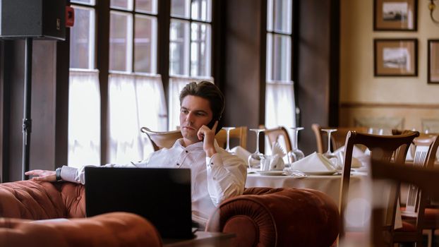 Portrait of goodlooking man sitting at table at home with laptop computer talking on the phone