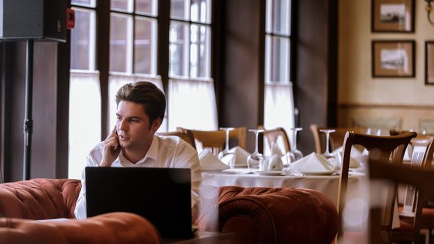 Portrait of goodlooking man sitting at table at home with laptop computer talking on the phone