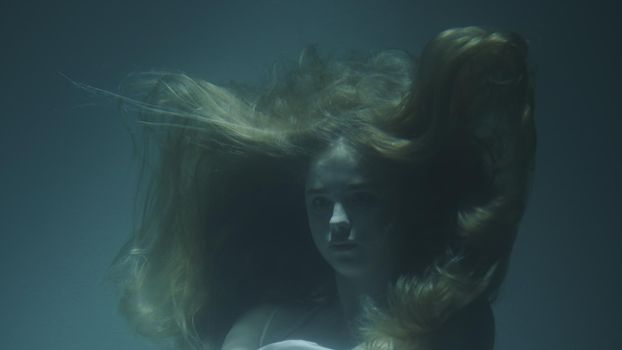 Close-up of a beautiful girl with red hair in a white dress froze under water in 4k