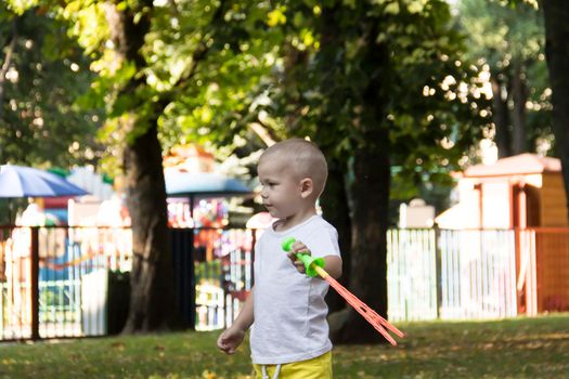 A little bald boy in yellow shorts is having fun blowing soap bubbles in an amusement park.