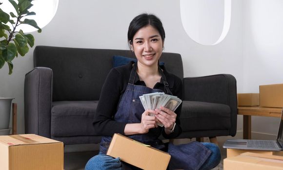 Young woman freelancer working and holding money with cardboard box at home - SME business online and delivery concept.