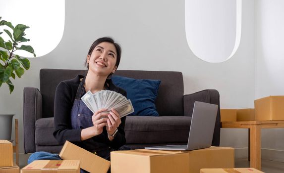 Young woman freelancer working and holding money with cardboard box at home - SME business online and delivery concept.