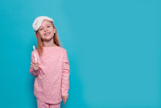close-up of a blonde, caucasian girl in pink pajamas, a unicorn-like sleep mask. The child shows an toothbrush in the hand. Blue background, copy space. The concept of children's health, good sleep