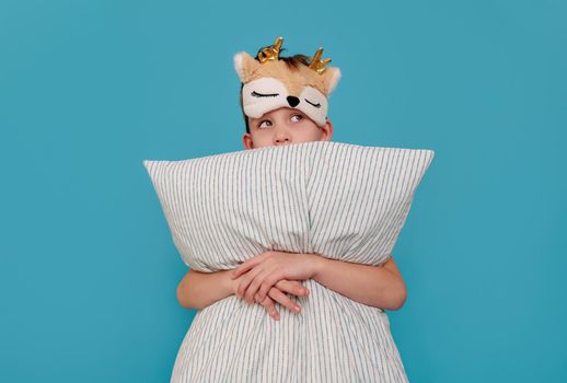the cunning boy does not want to sleep, hugs the pillow and looks away. Child on a blue background with copy space. The concept of a naughty child, parenting, insomnia, health