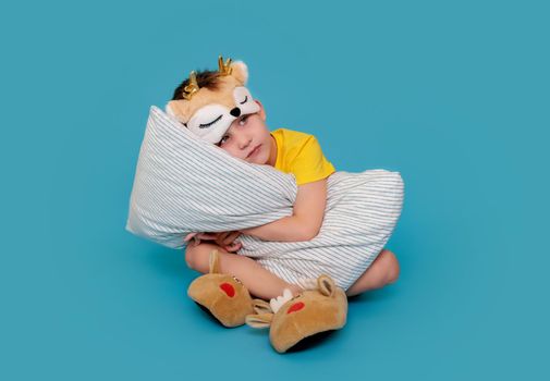 The little boy is tired and wants to sleep, hugs the pillow. The child is dressed in pajamas and a sleep mask, on his feet are funny slippers like deer. poster with blue background and copy space