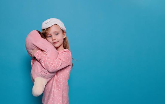 close-up of a blonde, caucasian girl in pink pajamas and a unicorn-like sleep mask. The child hugs a plush big toy. Blue background, copy space. The concept of children's health, good sleep