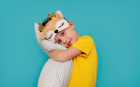 Banner, portrait of a sad boy on a blue background with copy paste. The child hugs the pillow. The boy is wearing yellow pajamas and a sleep mask, deer. Concept, rest, tired child, insomnia