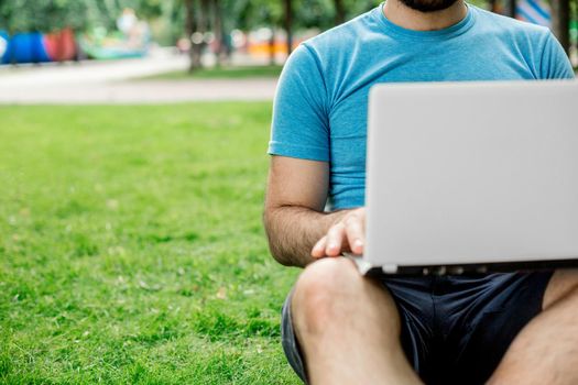 Young man using and typing laptop computer in summer grass. Freelancer working in outdoor park