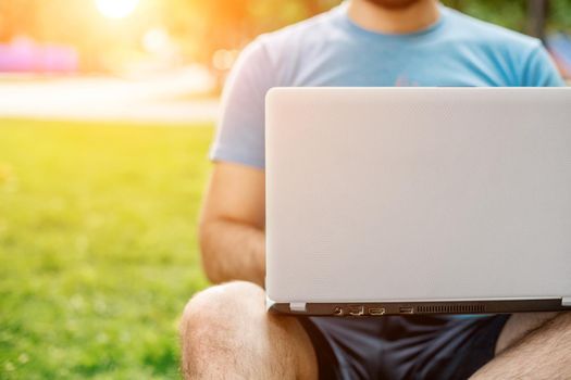 Young man using and typing laptop computer in summer grass. Freelancer working in outdoor park. Sun flare