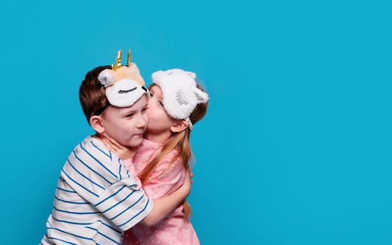 close-up of a blonde European girl in pink pajamas and a unicorn sleep mask. A boy in striped pajamas and a sleep mask. Children hugging, kissing and laughing on a blue background, copy paste