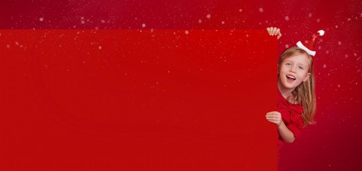 Long banner, red background with copy space. Merry Christmas and Happy New Year. Little blonde, caucasian woman in a santa claus hat, laughs happily, peeks out from behind a large piece of paper